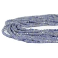 Drobne koraliki w kształcie fasetowanych kostek tanzanitu, w rozmiarze ok. 3x3mm, w odcieniach koloru niebiesko fioletowego. Nawleczone na różowe sznurki, ułożone w zwiniętej kiści, na białym tle. Widok z perspektywy bocznej.