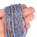 Drobne koraliki w kształcie fasetowanych kostek tanzanitu, w rozmiarze ok. 3x3mm, w odcieniach koloru niebiesko fioletowego. Nawleczone na różowe sznurki, prezentowane na dłoni, na białym tle.