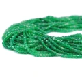 Drobne koraliki w kształcie fasetowanych kostek onyksu zielonego, w rozmiarze ok. 3x3mm. Nawleczone na zielone sznurki, ułożone w zwiniętej kiści, na białym tle. Widok z perspektywy bocznej.