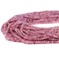 Drobne koraliki w kształcie fasetowanych kostek różowego turmalinu, w rozmiarze ok. 3x3mm. Nawleczone na różowe sznurki, ułożone w zwiniętej kiści, na białym tle. Widok z perspektywy bocznej.