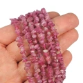 Kamienie w formie drobnych, nieregularnych bryłek różowego turmalinu, w rozmiarze ok. 4-8mm. Prezentowane na dłoni, na białym tle.