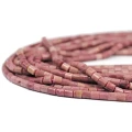 Kamienie w formie gładkich walców z różowego rodonitu, o wymiarach ok. 4 × 4,5 mm. Nawleczone na różowe sznurki. Widok z perspektywy bocznej – leżą w zwiniętej kiści na białym tle.