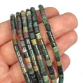 Kamienie w formie gładkich walców z agatu indyjskiego, w odcieniach zieleni i brązu, o wymiarach ok. 4 × 4,5 mm. Nawleczone na zielone sznurki.  Prezentowane na dłoni, na białym tle.