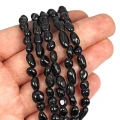 Kamienie w formie nieregularnych gładkich bryłek czarnego turmalinu, w rozmiarze ok. 5-10x4-6mm. Nawleczone na czarne sznurki. Prezentowane na dłoni, na białym tle.